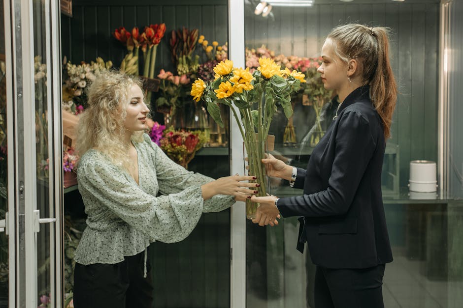 A florist handing over a bouquet of bright yellow and orange sunflowers to a customer inside a flower shop. The customer, a woman with curly blonde hair wearing a light green patterned blouse, is reac