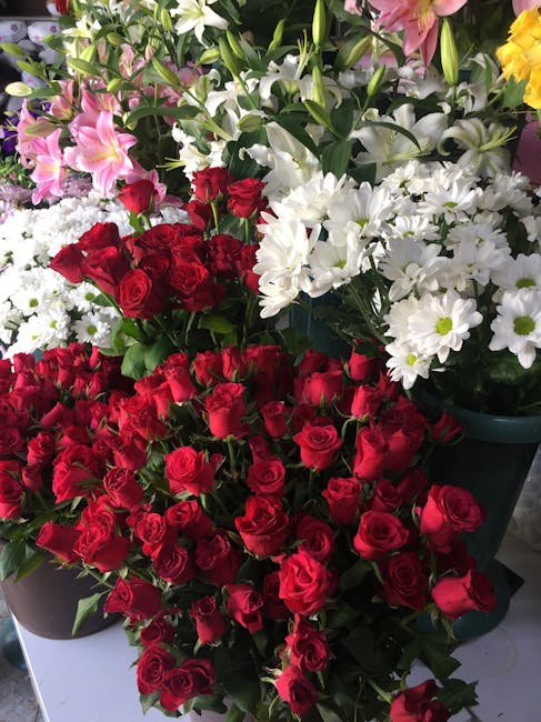 A vibrant display of fresh floral arrangements featuring lush red roses in the foreground, accompanied by white daisies with yellow centers, pink lilies with delicate petals, and yellow flowers in the