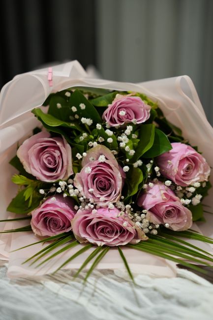 A bouquet of fresh pink roses with delicate, tightly layered petals, accompanied by small white baby's breath flowers and lush green foliage. The roses are arranged in a compact, rounded style, with g