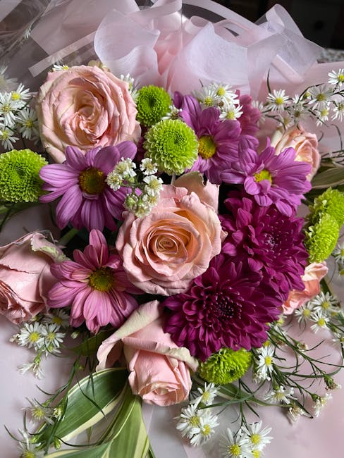 A vibrant floral bouquet featuring pale pink roses with soft, layered petals, purple daisies with bright yellow centers, and small white daisy-like flowers with yellow centers, all arranged with fresh
